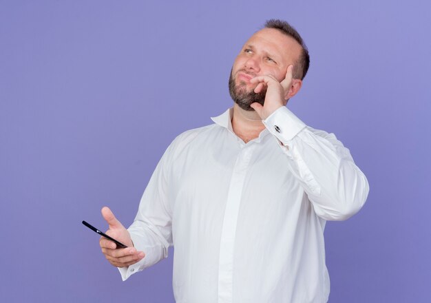 Hombre barbudo vestido con camisa blanca sosteniendo smartphone mirando hacia arriba con expresión pensativa de pie sobre la pared azul