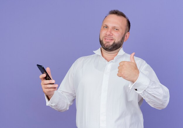 Hombre barbudo vestido con camisa blanca sosteniendo smartphone mirando sonriendo mostrando Thumbs up de pie sobre la pared azul