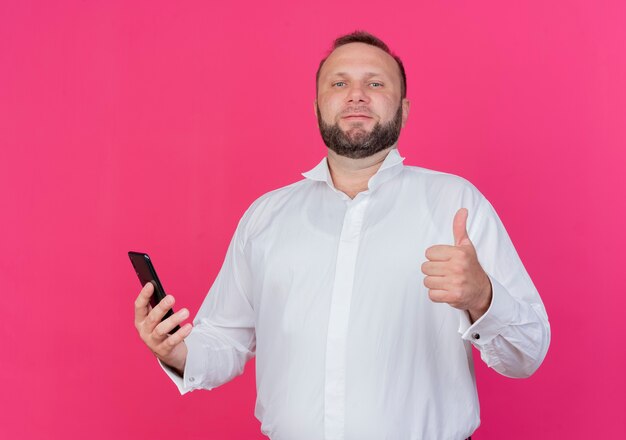 Hombre barbudo vestido con camisa blanca sosteniendo smartphone mostrando los pulgares para arriba de pie sobre la pared rosa