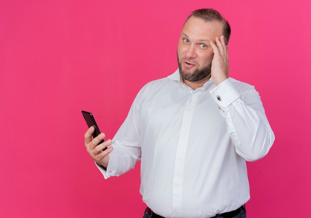 Hombre barbudo vestido con camisa blanca sosteniendo smartphone sonriendo de pie sobre la pared rosa