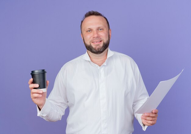 Hombre barbudo vestido con camisa blanca sosteniendo una taza de café y una página en blanco mirando sonriente de pie sobre la pared azul