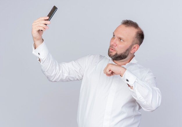 Hombre barbudo vestido con camisa blanca sosteniendo teléfono inteligente con videollamada mirando la pantalla con cara seria de pie sobre la pared blanca