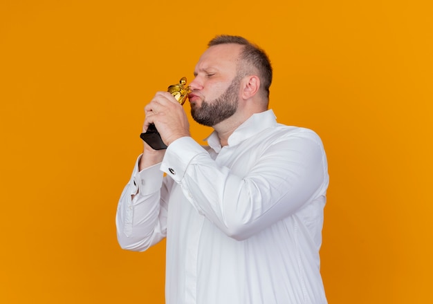 Hombre barbudo vestido con camisa blanca sosteniendo trofeo besándolo feliz y emocionado de pie sobre la pared naranja