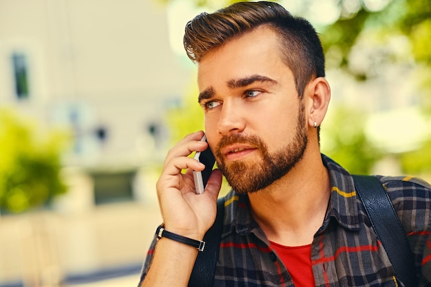 Hombre barbudo vestido con una camisa de lana habla por un teléfono inteligente en un parque. De cerca.