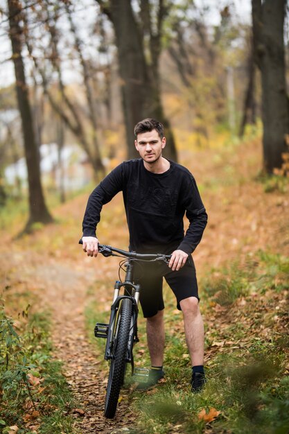 Hombre bastante atlético joven que se coloca con la bicicleta en parque colorido del otoño. Temporada de otoño. Ciclista masculino en el camino con hojas caídas