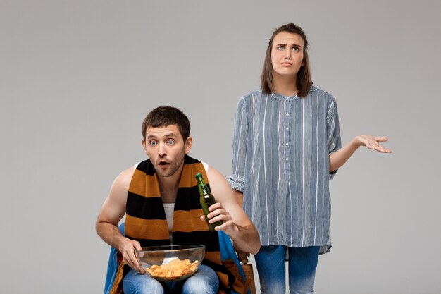 Hombre bebiendo cerveza, viendo fútbol y molesta mujer de pie detrás