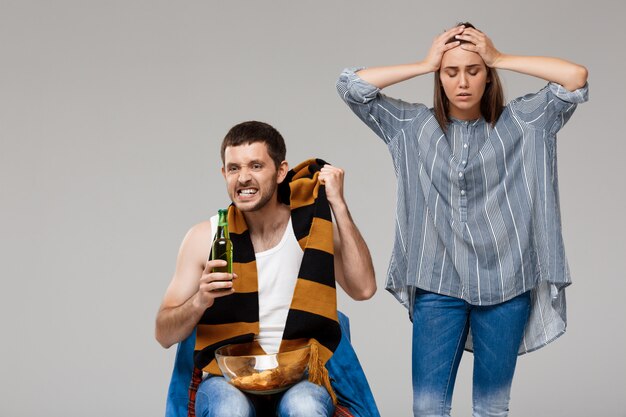 Hombre bebiendo cerveza, viendo fútbol y molesta mujer de pie detrás