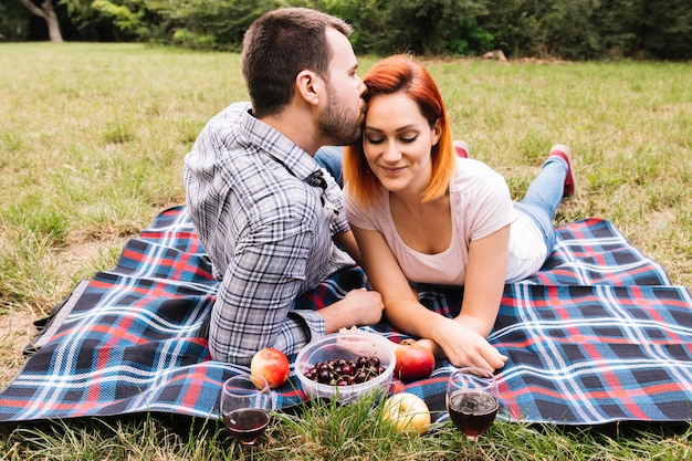 Hombre besando a su novia tendida en una manta sobre hierba verde con frutas