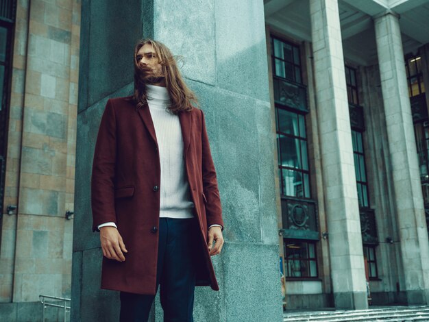Hombre bien vestido confiado hermoso con la barba que presenta al aire libre que mira lejos. Modelo masculino rico de moda en abrigo de invierno rojo y suéter blanco