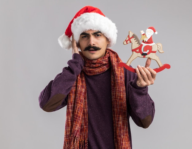 Hombre con bigote joven vestido con gorro de Papá Noel de Navidad con bufanda alrededor de su cuello sosteniendo un juguete de Navidad mirando confundido