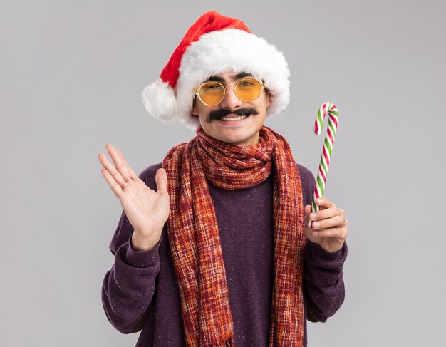 Hombre bigotudo feliz con gorro de santa de navidad y gafas amarillas con bufanda alrededor de su cuello sosteniendo bastón de caramelo sonriendo alegremente de pie sobre la pared blanca