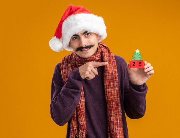 Hombre bigotudo con gorro de navidad de santa con bufanda caliente alrededor de su cuello sosteniendo cubos de juguete con fecha veinticinco apuntando con el dedo índice a los cubos sonriendo de pie sobre la pared naranja