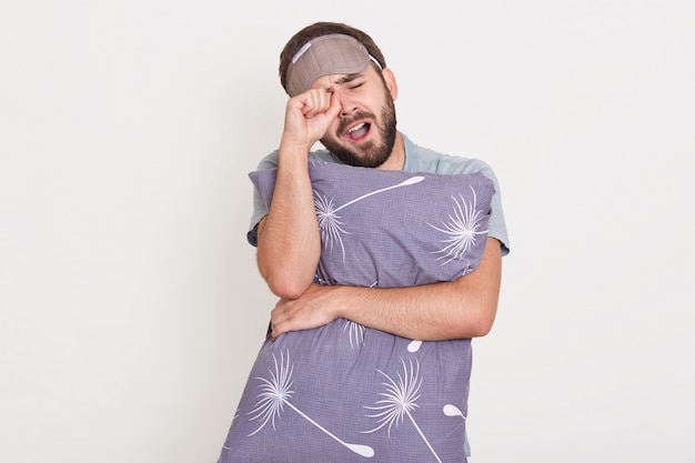 Hombre bostezo sin afeitar posando en el interior, frotándose los ojos, manteniendo la boca abierta, sosteniendo la almohada gris en las manos, chico despierto por la mañana