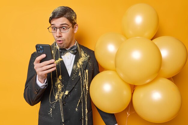 Un hombre brunet sorprendido mira fijamente la pantalla del teléfono inteligente y lee noticias increíbles vestido formalmente pasa tiempo libre en la fiesta sostiene un montón de globos inflados aislados sobre un fondo amarillo vívido Festividad