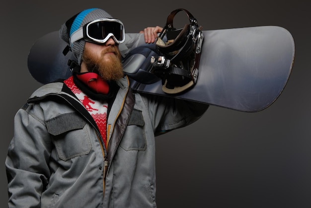 Un hombre brutal con barba roja usando un equipo completo sosteniendo una tabla de snowboard en su hombro, aislado en un fondo oscuro.