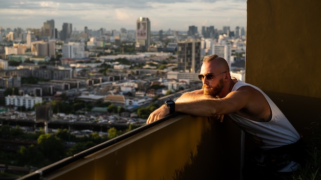 Hombre brutal barbudo guapo caucásico disfrutando de la puesta de sol y vista de la ciudad de Bangkok desde el piso alto