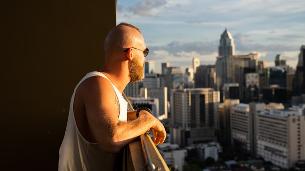 Hombre brutal barbudo guapo caucásico disfrutando de la puesta de sol y vista de la ciudad de Bangkok desde el piso alto