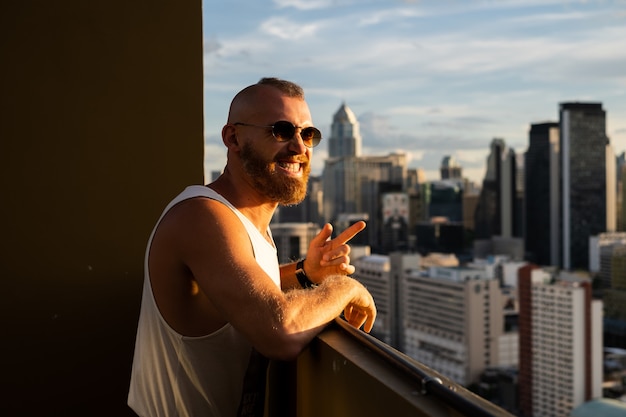 Hombre brutal barbudo guapo caucásico disfrutando de la puesta de sol y vista de la ciudad de Bangkok desde el piso alto