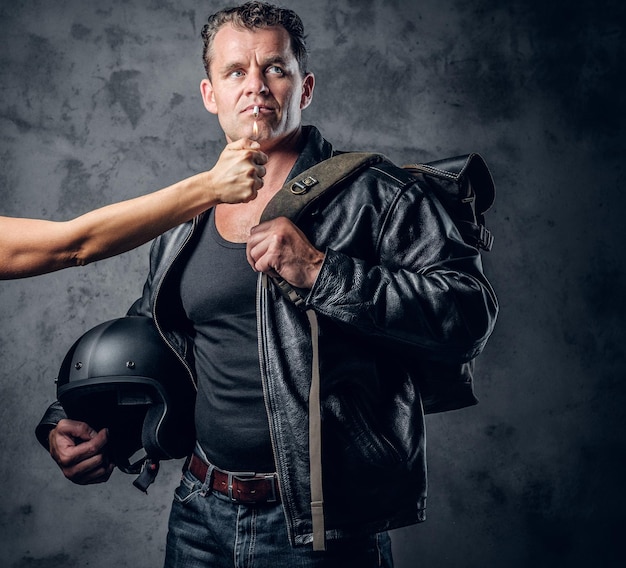 Hombre brutal con chaqueta de cuero sostiene casco de motocicleta y enciende un cigarrillo del brazo de la mujer.