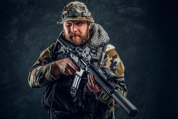 Hombre brutal con uniforme militar camuflado sosteniendo un rifle de asalto. Foto de estudio contra una pared de textura oscura