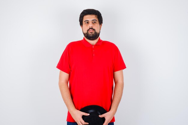 Hombre de cabello oscuro con camiseta roja sosteniendo sombrero y mirando tranquilo, vista frontal.