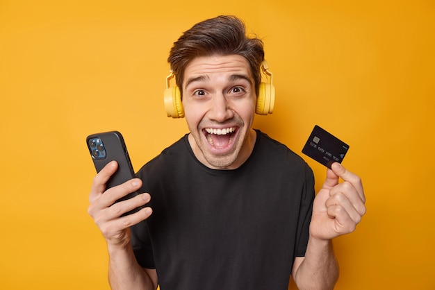 Un hombre de cabello oscuro lleno de alegría usa una aplicación de teléfono móvil para pagar en línea, tiene una tarjeta de crédito, hace compras, ordena algo a través de un celular, usa auriculares en las orejas aisladas sobre una pared amarilla del estudio