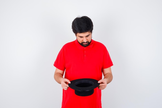 Hombre de cabello oscuro mirando dentro de su sombrero en la vista frontal de la camiseta roja.