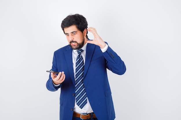 Hombre de cabello oscuro mirando el teléfono con camisa blanca, chaqueta azul oscuro, corbata y aspecto agresivo, vista frontal.