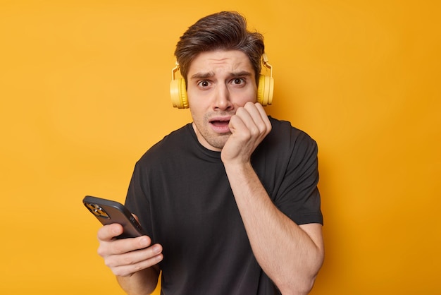 El hombre de cabello oscuro preocupado se siente nervioso, se muerde las uñas y escucha música a través de los auriculares, usa una camiseta negra informal aislada sobre fondo amarillo. Concepto de tecnología y entretenimiento de la gente.