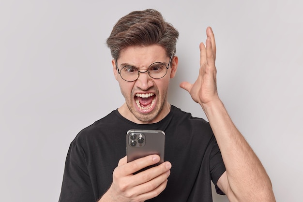 Un hombre cabreado y molesto grita airadamente y mantiene la palma de la mano mirando fijamente al teléfono inteligente indignado después de que una conversación áspera lleva anteojos redondos, una camiseta negra informal aislada sobre fondo blanco