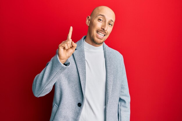 Hombre calvo con barba con chaqueta de negocios sonriendo con una idea o pregunta señalando con el dedo hacia arriba con cara feliz, número uno