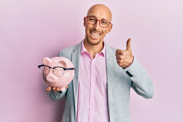 Hombre calvo con barba sosteniendo alcancía con gafas sonriendo feliz y positivo, pulgar arriba haciendo excelente y señal de aprobación