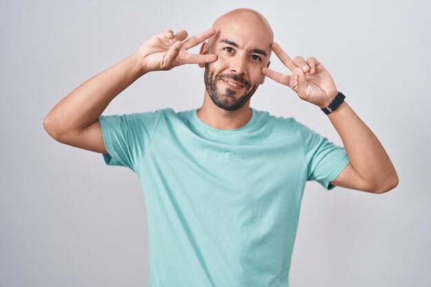 Hombre calvo de mediana edad parado sobre fondo blanco haciendo símbolo de paz con los dedos sobre la cara, sonriendo alegre mostrando victoria