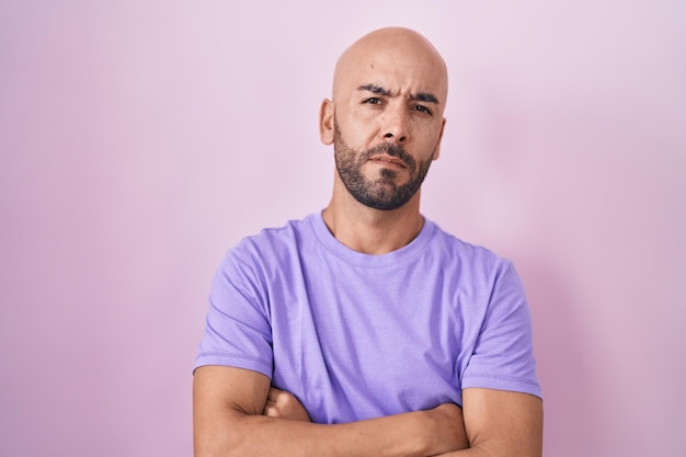 Hombre calvo de mediana edad parado sobre un fondo rosado escéptico y nervioso desaprobando la expresión en la cara con los brazos cruzados persona negativa