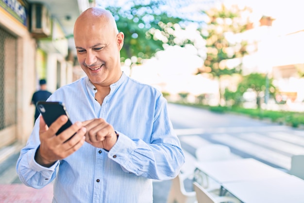 Hombre calvo de mediana edad sonriendo feliz usando un smartphone en la ciudad.