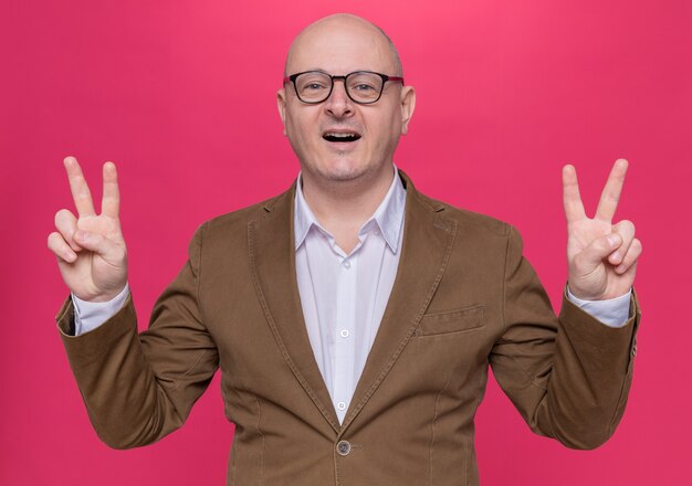 Hombre calvo de mediana edad en traje con gafas mirando al frente feliz y alegre sonriendo ampliamente mostrando v-sign de pie sobre la pared rosa