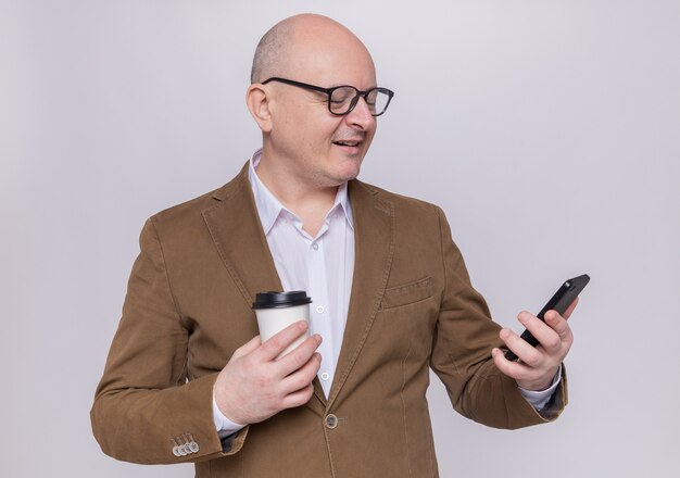 Hombre Calvo de mediana edad en traje con gafas mirando la pantalla de su teléfono móvil sosteniendo un vaso de papel sonriendo confiado de pie sobre la pared blanca