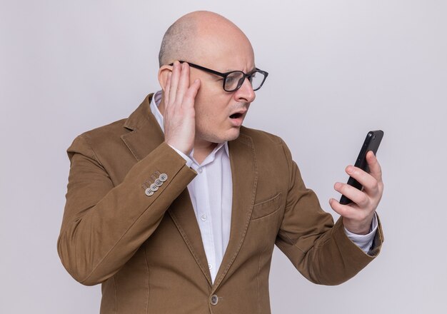 Hombre Calvo de mediana edad en traje con gafas mirando su teléfono móvil confundido