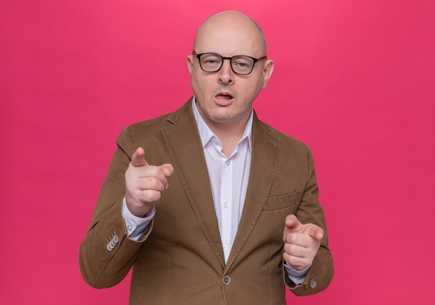 Hombre Calvo de mediana edad en traje con gafas sonriendo confiado apuntando con los dedos índices en la parte delantera de pie sobre la pared rosa