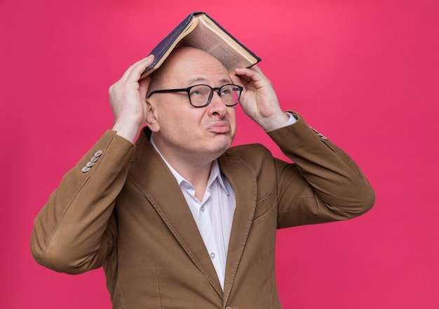 Hombre Calvo de mediana edad en traje con gafas sosteniendo un libro sobre su cabeza mirando confundido