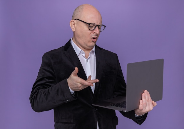 Hombre Calvo de mediana edad en traje con gafas sosteniendo portátil mirando la pantalla confundido y disgustado de pie sobre la pared púrpura