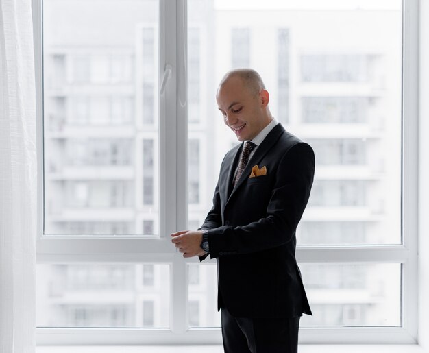 Un hombre calvo y sonriente se viste cerca de la ventana con el elegante traje antes de una importante reunión de negocios