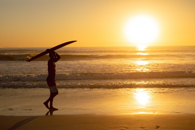 Hombre caminando con tabla de surf sobre su cabeza en la playa