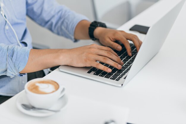 Hombre de camisa azul con portátil para trabajar, escribiendo en el teclado. Retrato interior de manos masculinas en la computadora y una taza de café en la mesa.