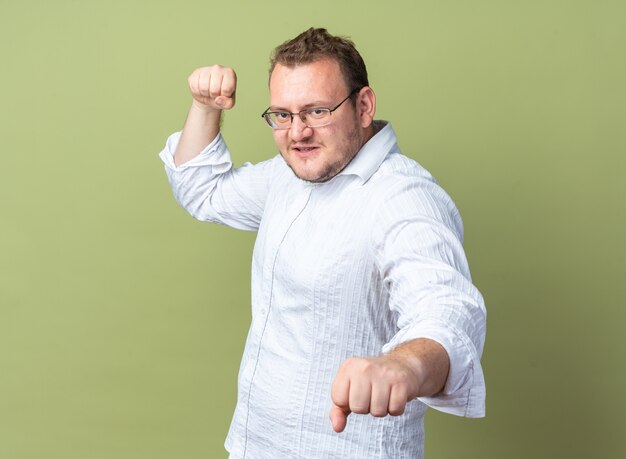 Hombre con camisa blanca con gafas mirando al frente con rostro serio apretando los puños de pie sobre la pared verde