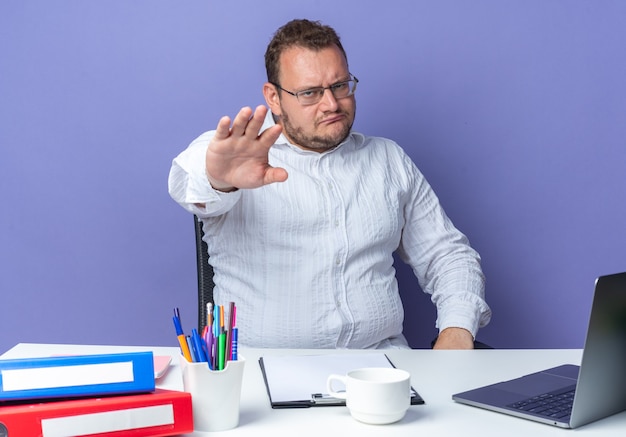 Hombre de camisa blanca con gafas mirando con el ceño fruncido haciendo gesto de parada con la mano sentada en la mesa con carpetas de oficina y portátil sobre fondo azul trabajando en oficina