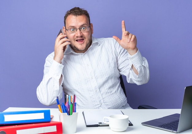Hombre con camisa blanca con gafas mirando sorprendido mientras habla por teléfono móvil sentado en la mesa con carpetas de oficina y portátil en azul