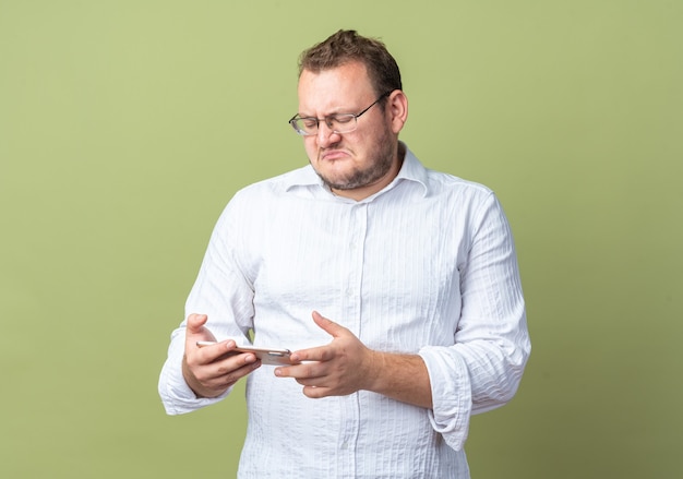 Hombre con camisa blanca con gafas sosteniendo smartphone mirándolo con expresión de decepción de pie sobre la pared verde