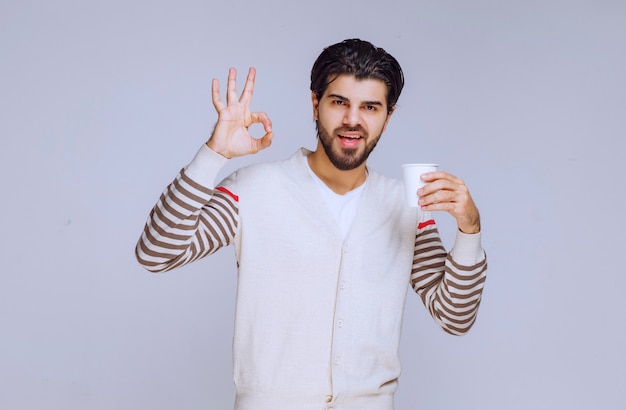 Hombre con camisa blanca sosteniendo una taza de café y haciendo un buen signo de mano.