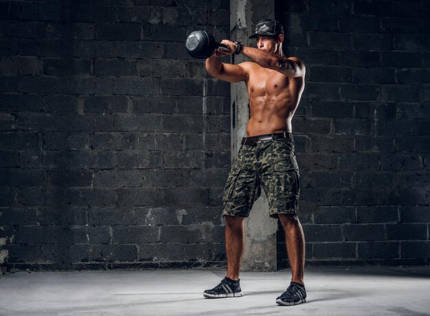 El hombre sin camisa enfocado con gorra está haciendo ejercicios con pesas en un estudio fotográfico oscuro.